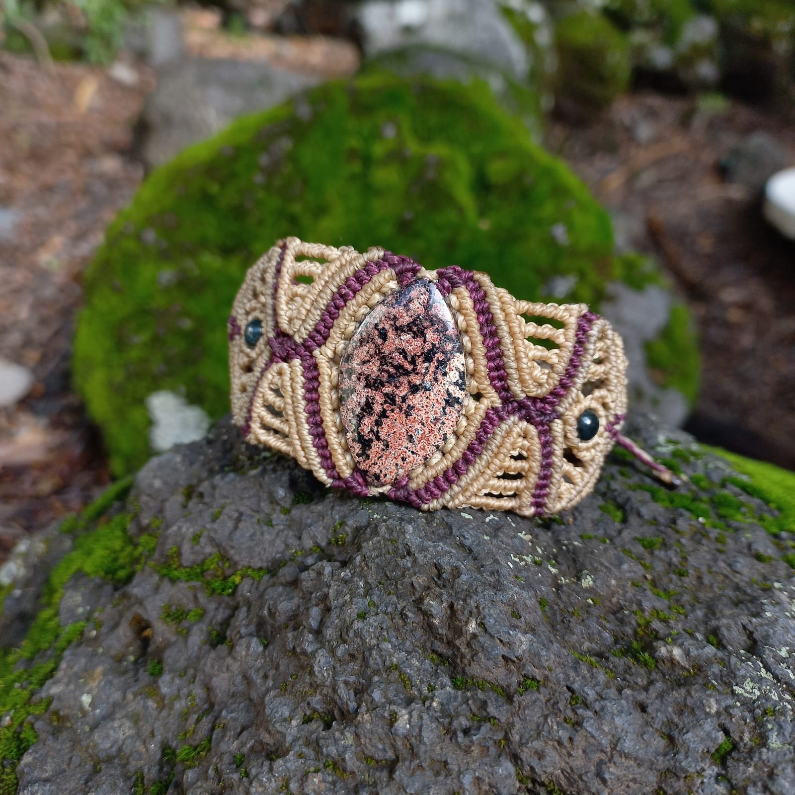 Pulsera de macramé beige y vino con cabujón de rodonita, sobre roca con musgo.